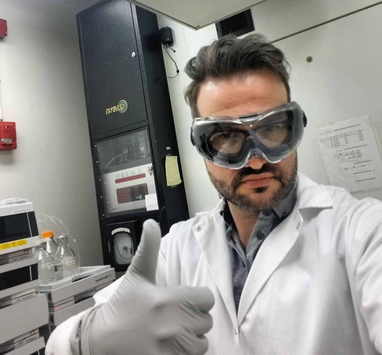 Erik Emilson wearing safety goggles and a white lab coat giving a thumbs up