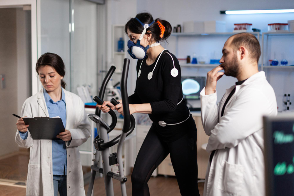 A woman on an elliptical with electrodes and a respiration monitoring mask on her, and with two researchers standing on each side of her monitoring her.
