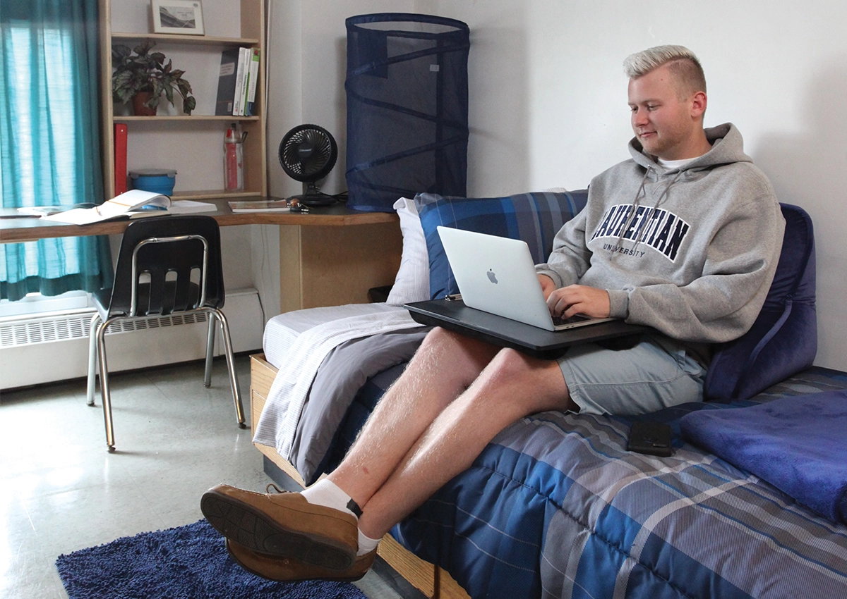 A Laurentian University student sitting on his bed in his residence room while on his laptop.