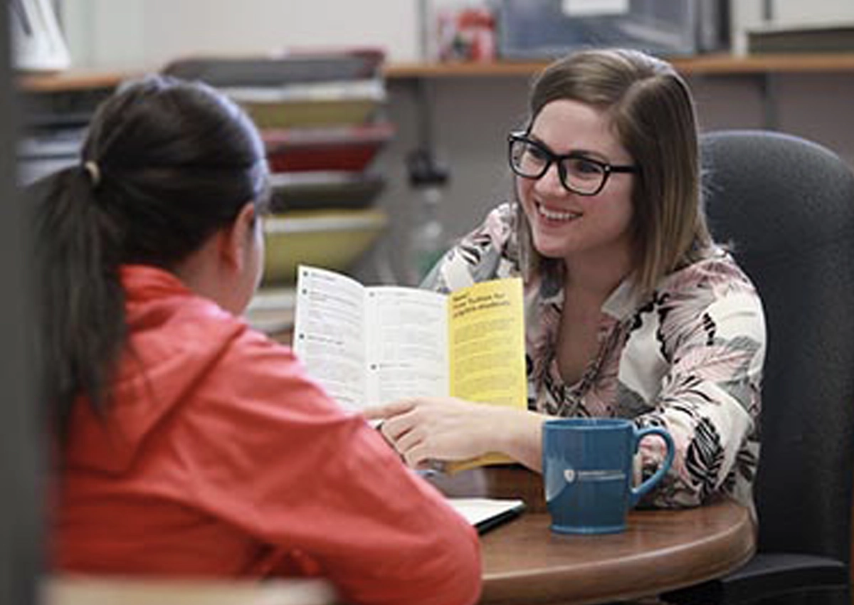 A woman smiling, showing a student an informative pamphlet.