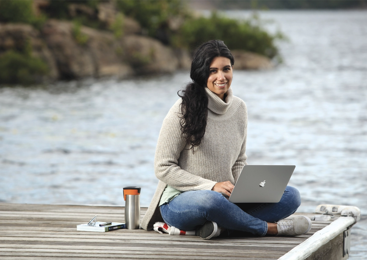 A woman sitting on a dock by the lake with her laptop.