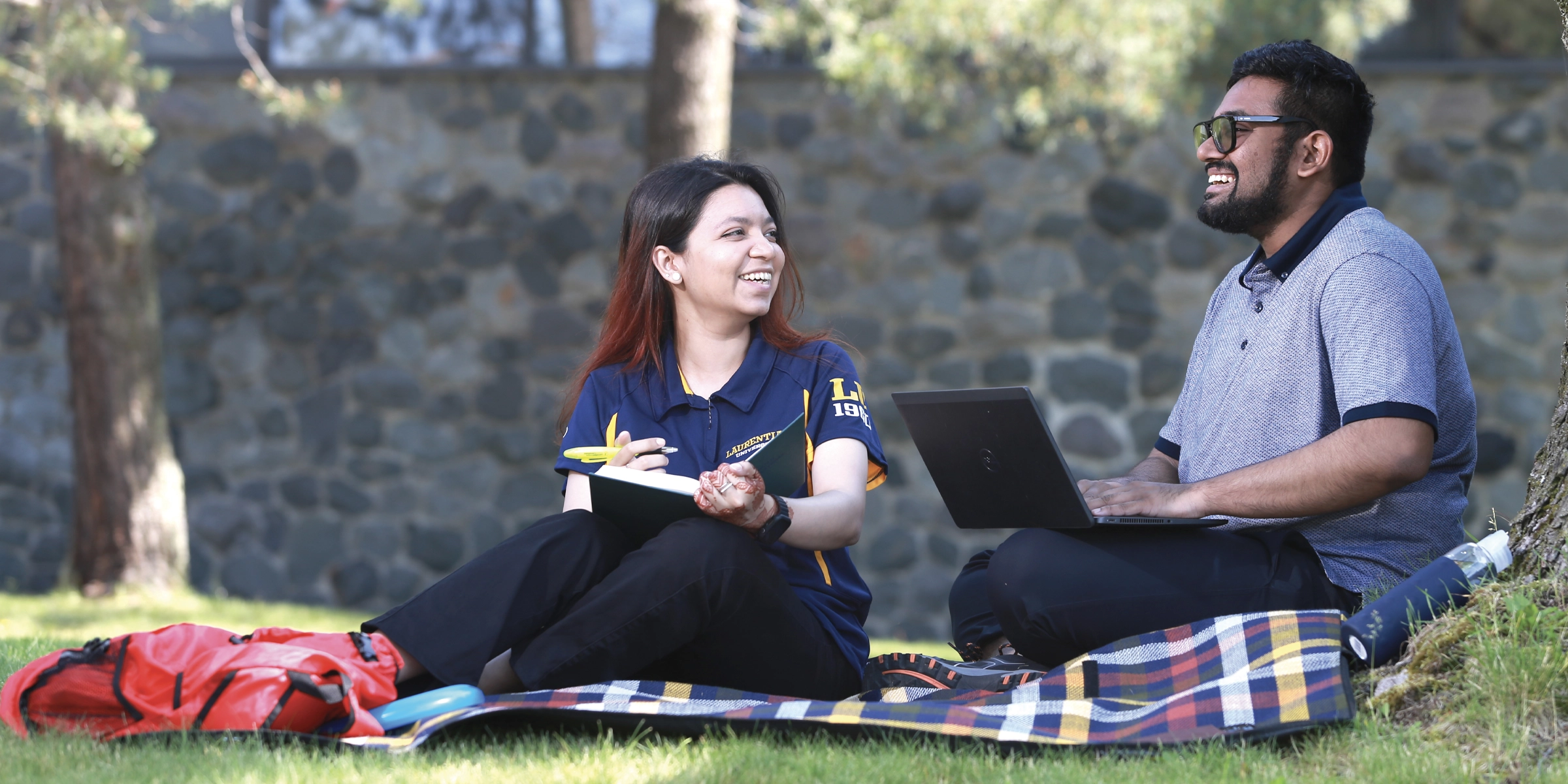 Two students sitting on the grass, working and laughing amongst each other.