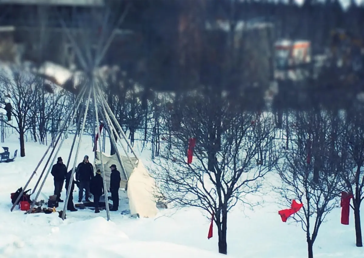 A snowy landscape with red dresses hanging from the trees, and people gathered in a tipi.