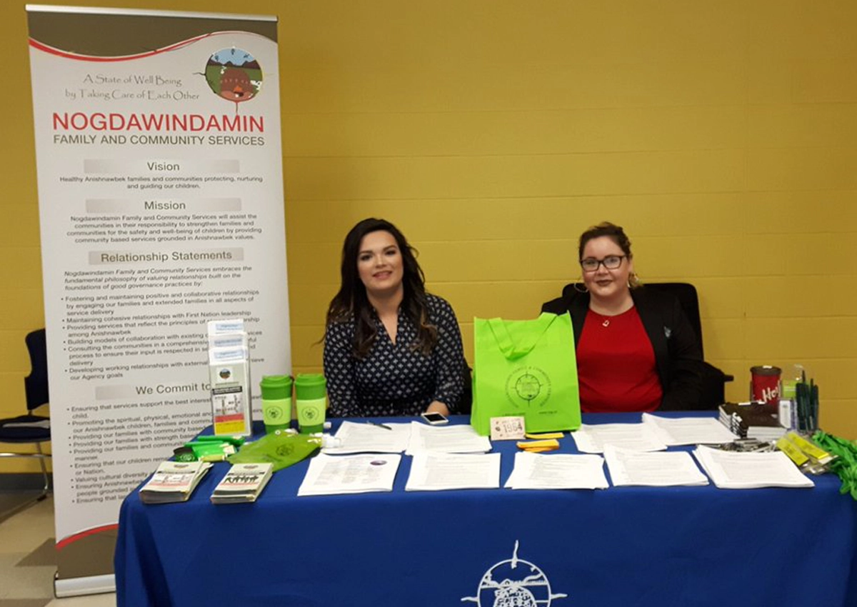 Two women sitting at a table for a services fair, with a banner that reads "Nogdawindamin Family and Community Services."