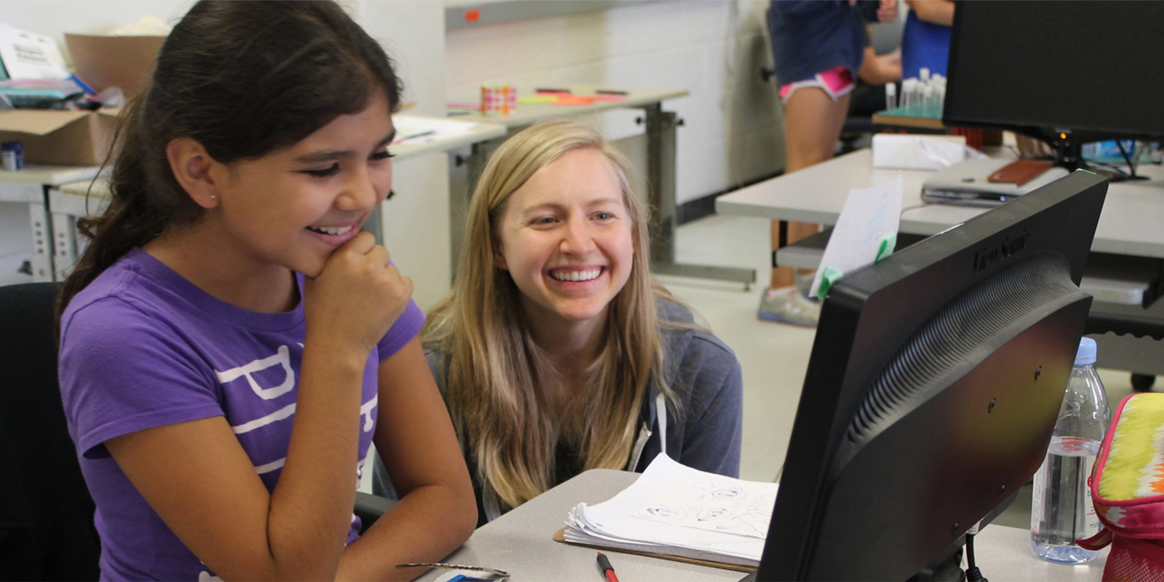 A young girl learning happily at a computer while her teacher sits next to her.