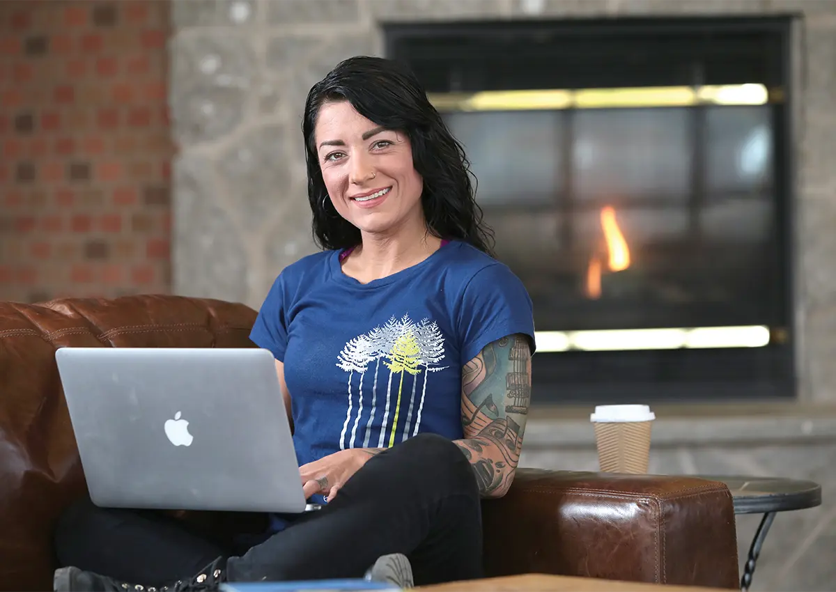 Woman sitting at couch with laptop open on lap looking at camera. 
