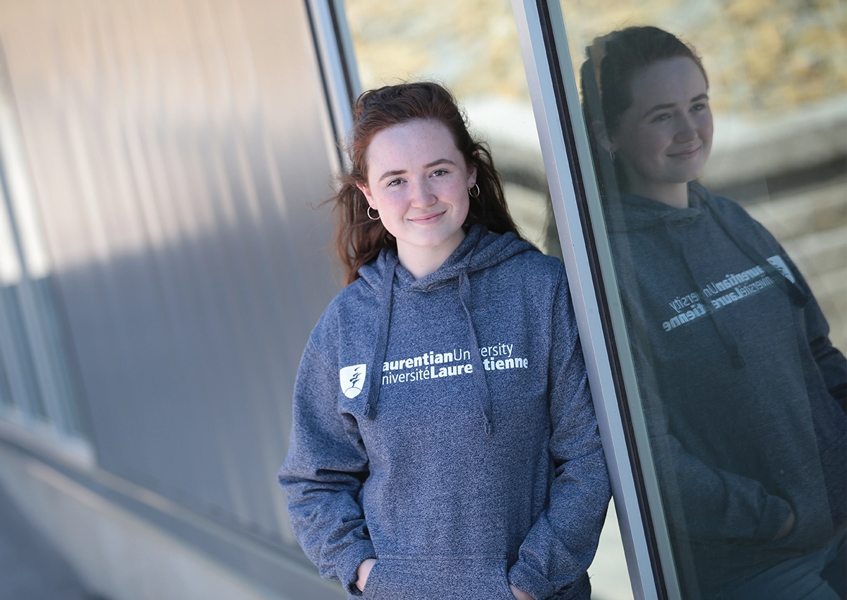A student leaning against a wall wearing a Laurentian University sweater looking towards the camera.