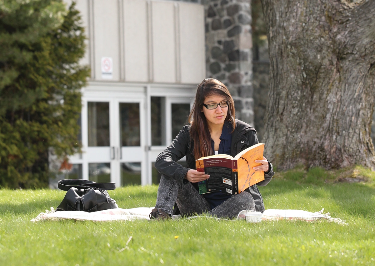 A Laurentian University student reading a book outside in front of the Arts Building.