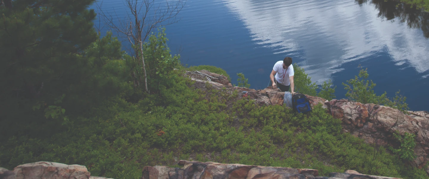 A student doing some mineral research on a large rock with a lake at his back.