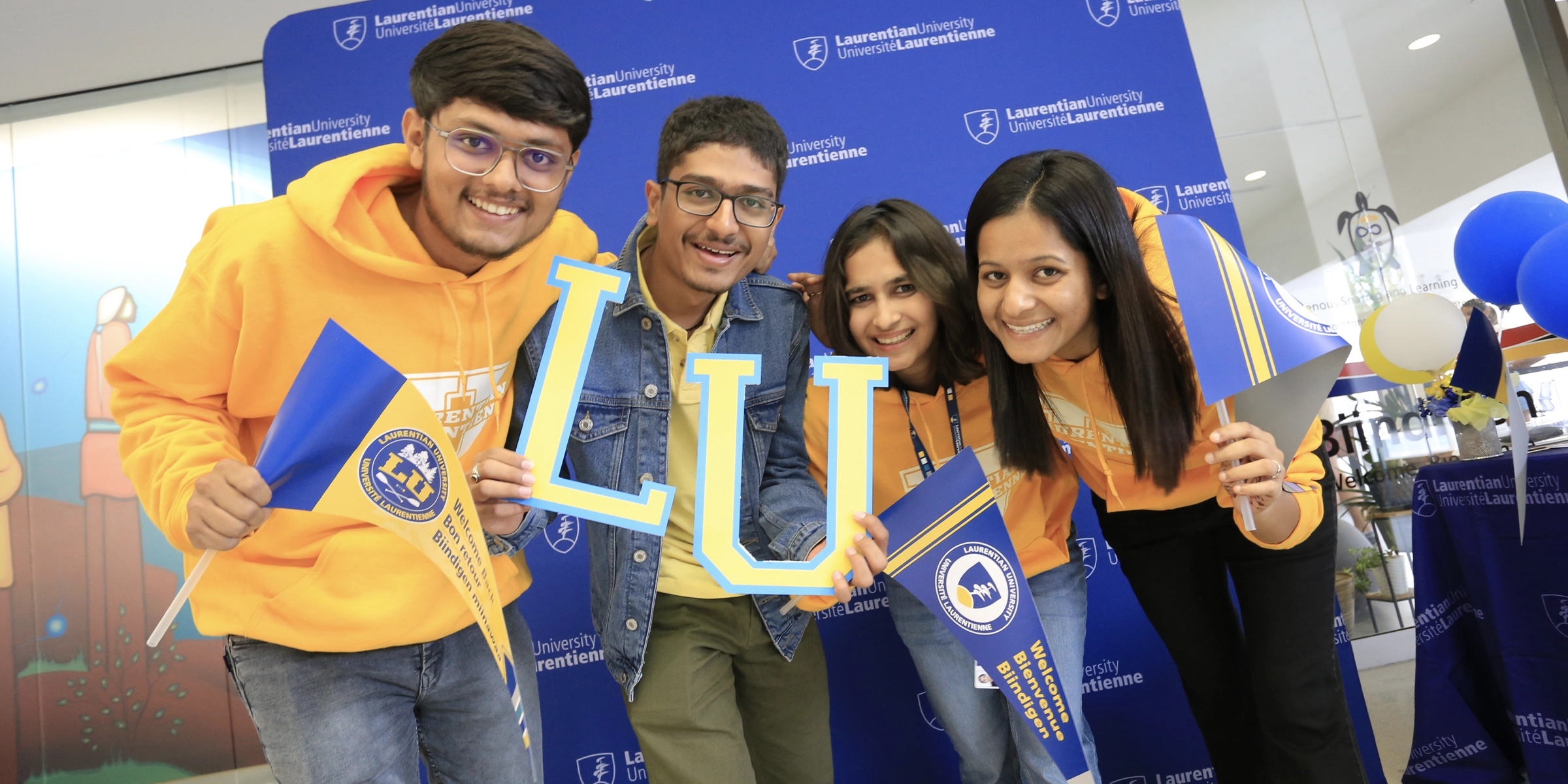 Four international students at Laurentian pose in front of a blue background, holding blue and yellow Laurentian flags.