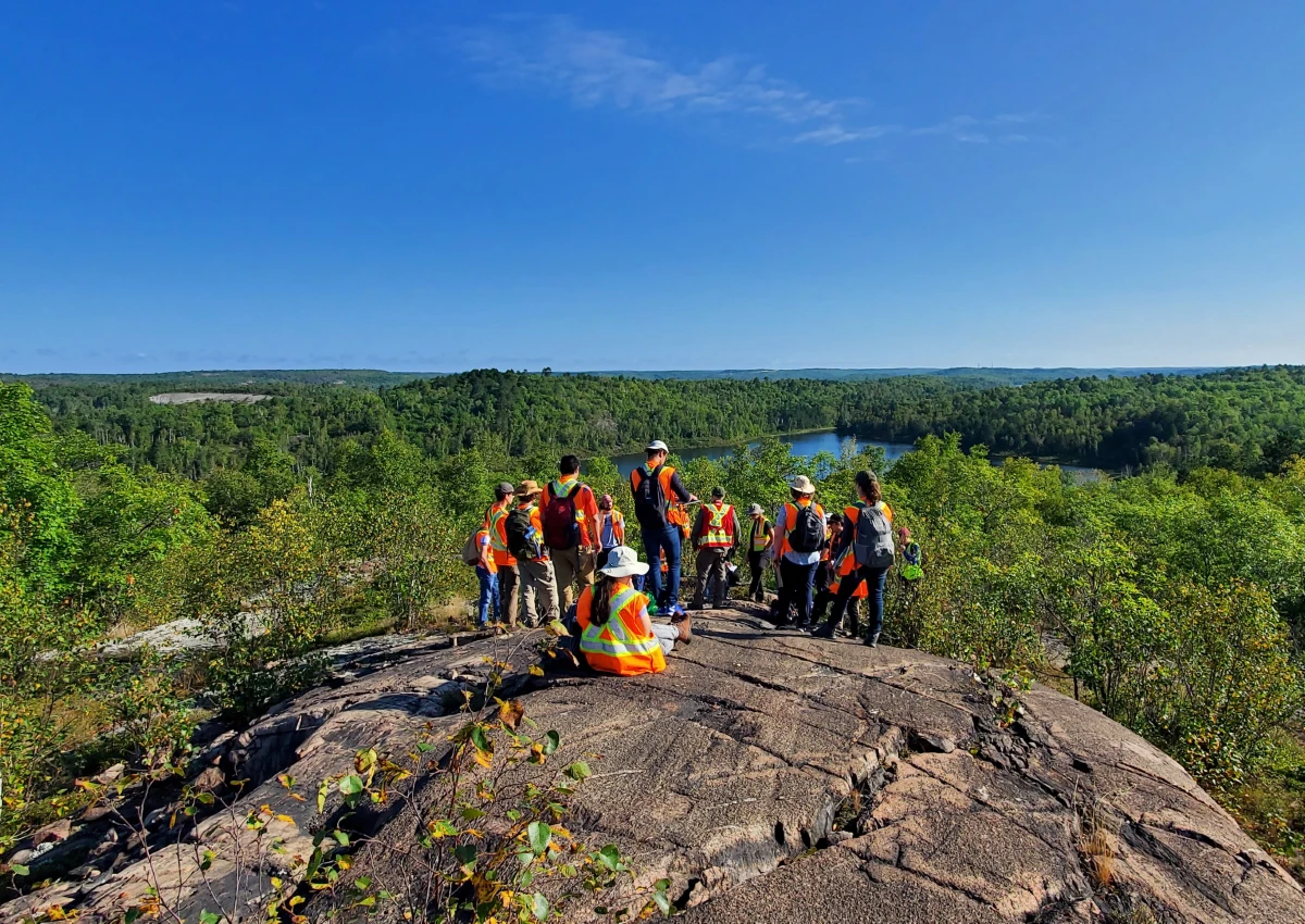 A group a students along with their prof are are top of a large rock overlooking a forest.