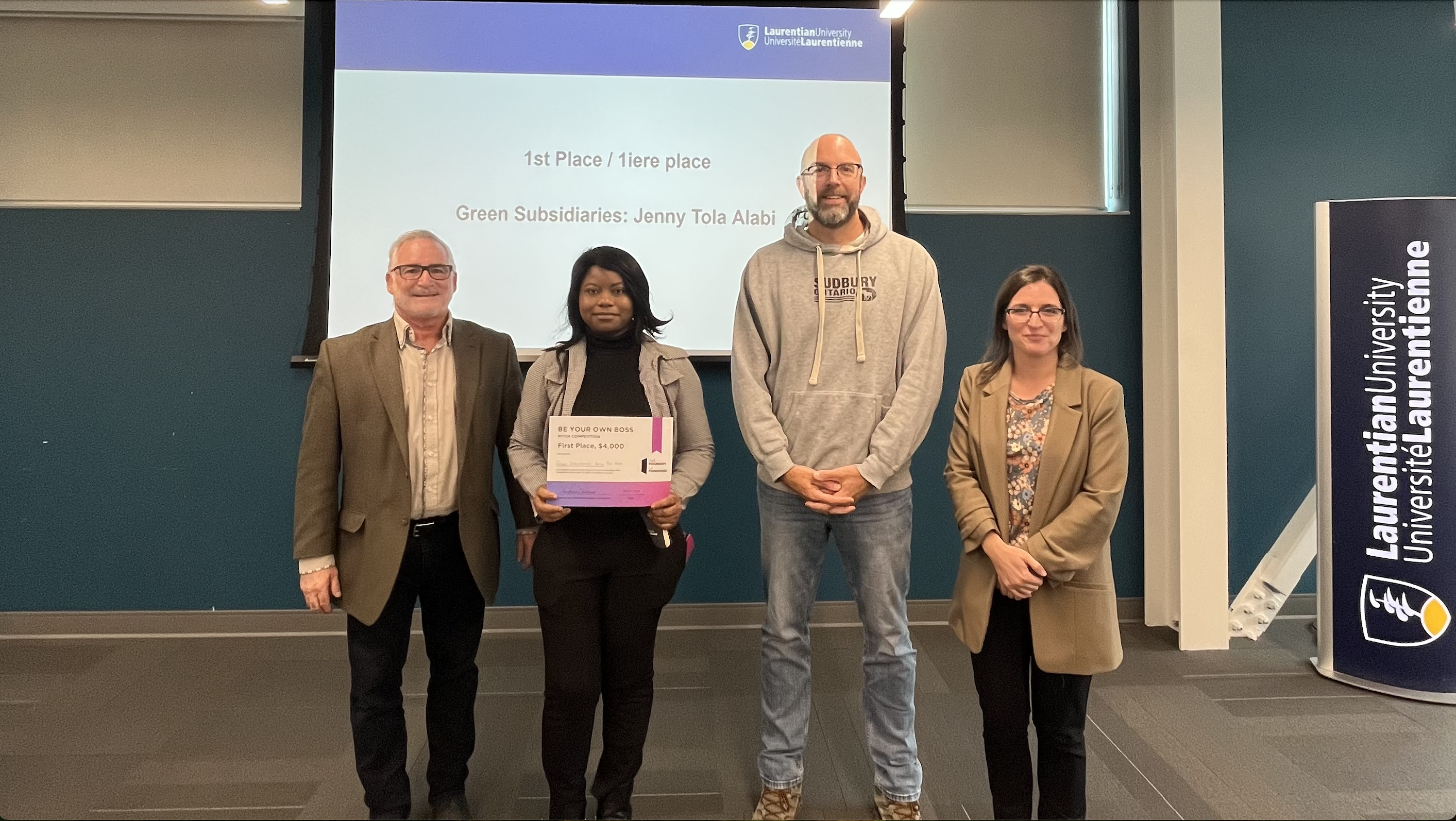 First-place BYOB winner Jenny Tola Alabi stands with three faculty members in front of a presentation screen that reads '1st Place / 1iere place - Green Subsidiaries: Jenny Tola Alabi