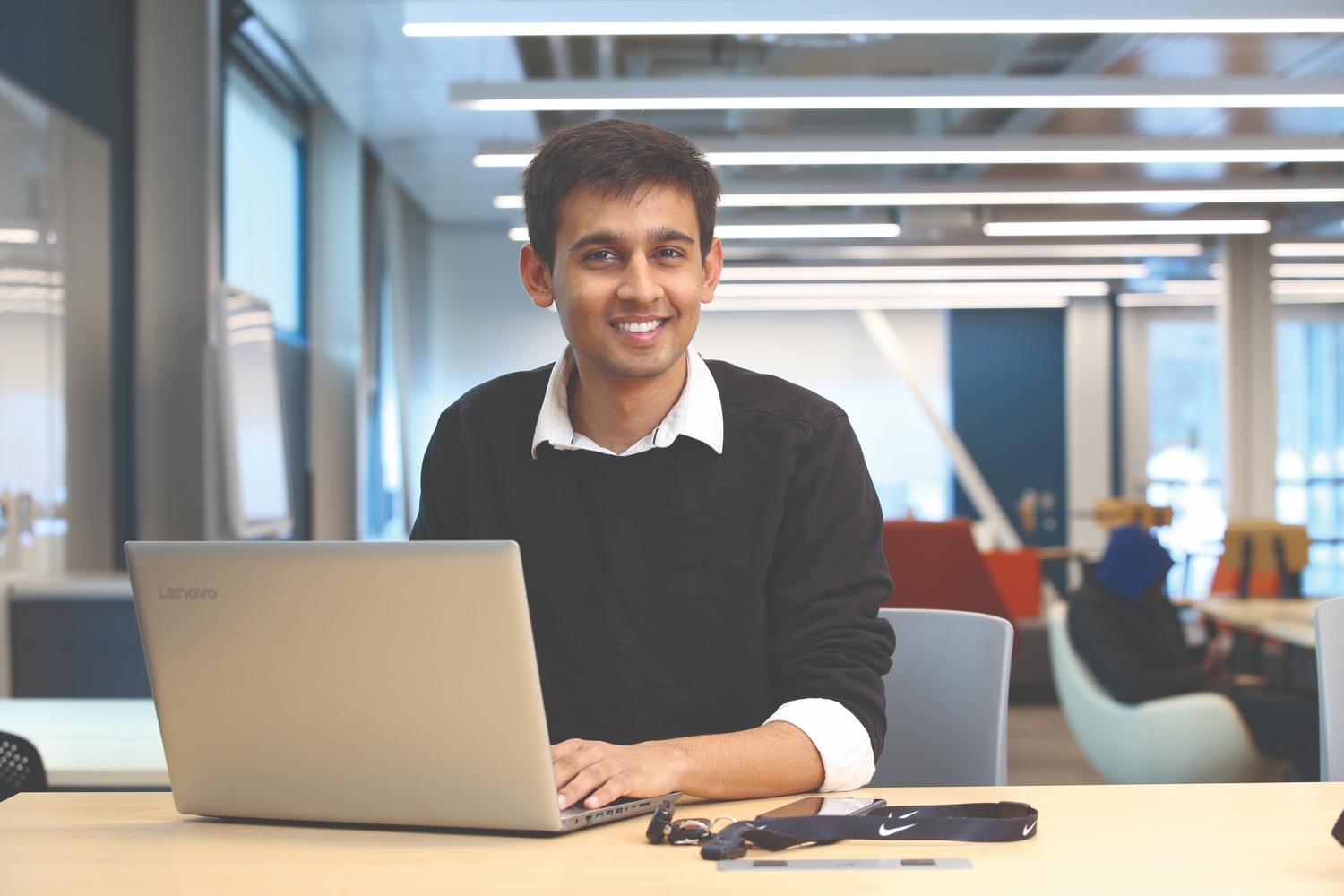 A male student is seated at a table smiling behind a laptop