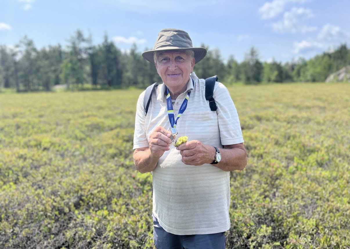 A wide-angle shot of Dr. Peter Beckett smiling at the camera in a vast, open wetland or peatland field. He is wearing a sun hat, a light-coloured striped polo shirt, and a lanyard, holding up a small sample of moss or lichen to examine.