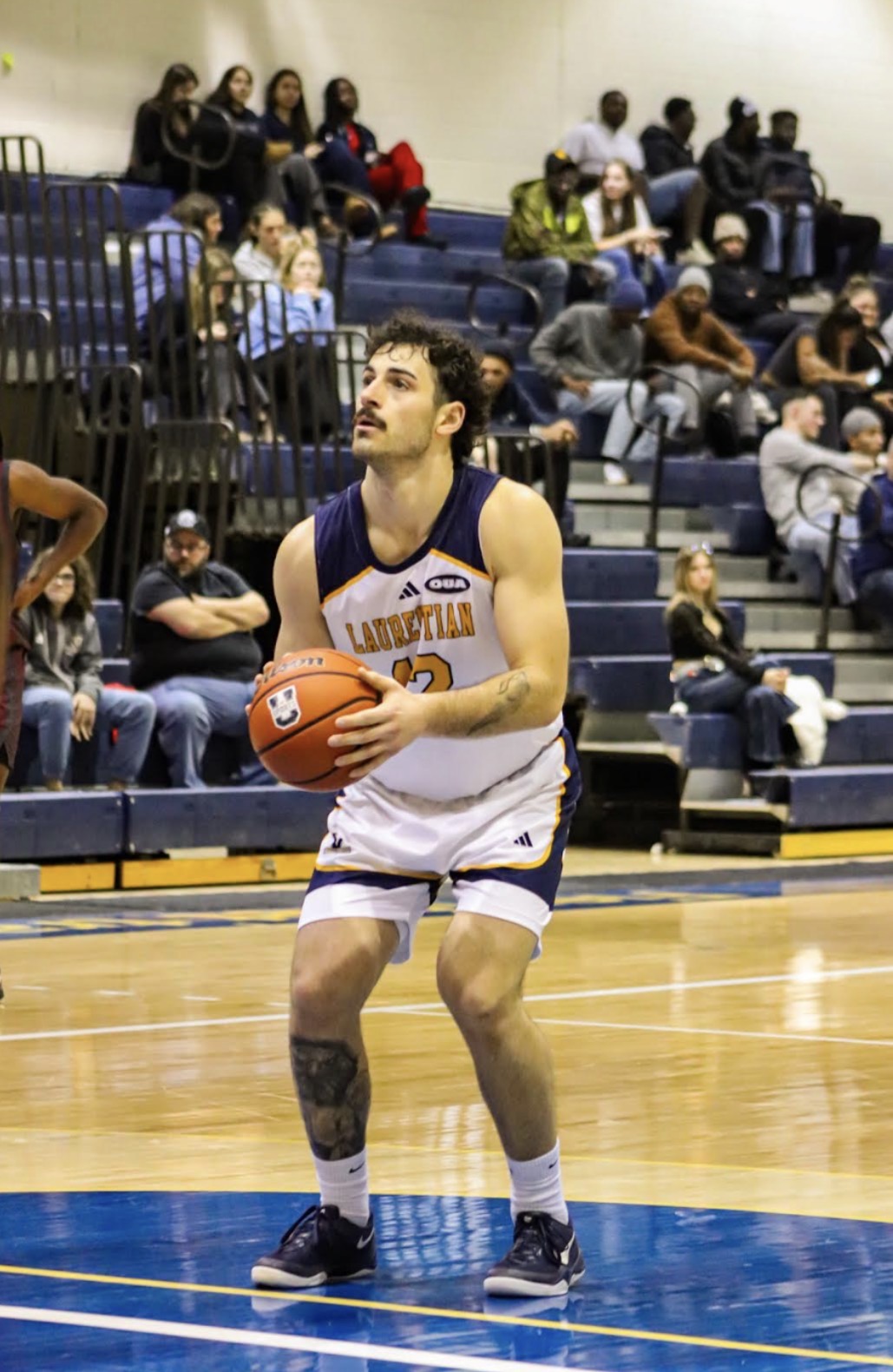 Evan Boudreau-Munro standing on a basketball court, looking up intently while preparing to take a shot. He is wearing a white Laurentian Voyageurs jersey with the number 12 and an Ontario University Athletics patch on the shoulder. He holds the basketball with both hands at chest level, with blue bleachers filled with spectators blurred in the background.