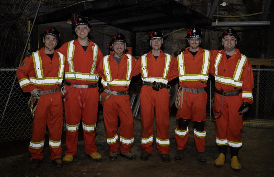 Six members of the Laurentian University Mine Rescue team—(from left to right) Félix Gervais, Martin Brunet, Aiden Alloy, Peter Guerra, Calvin Rapin, and David Langley—stand together in an underground mine. The students wear orange high-visibility coveralls, black hard hats, and safety gear.