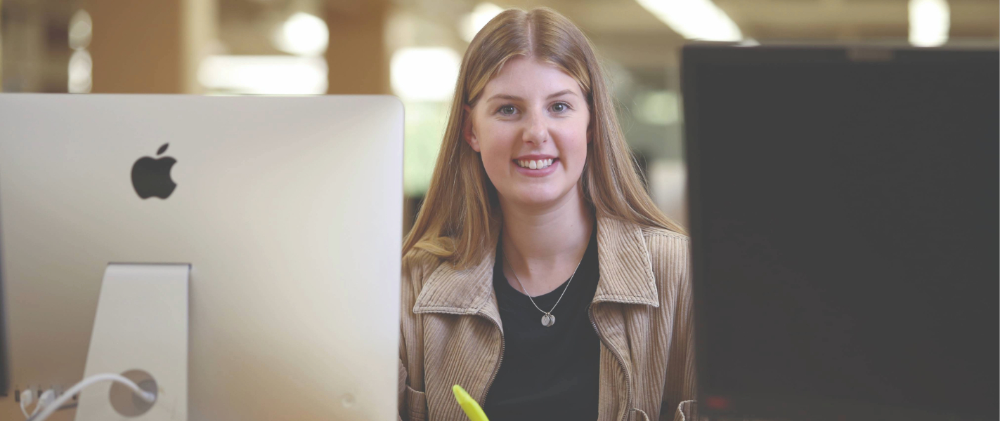 Student smiles towards camera, sitting behind a computer screen in the library