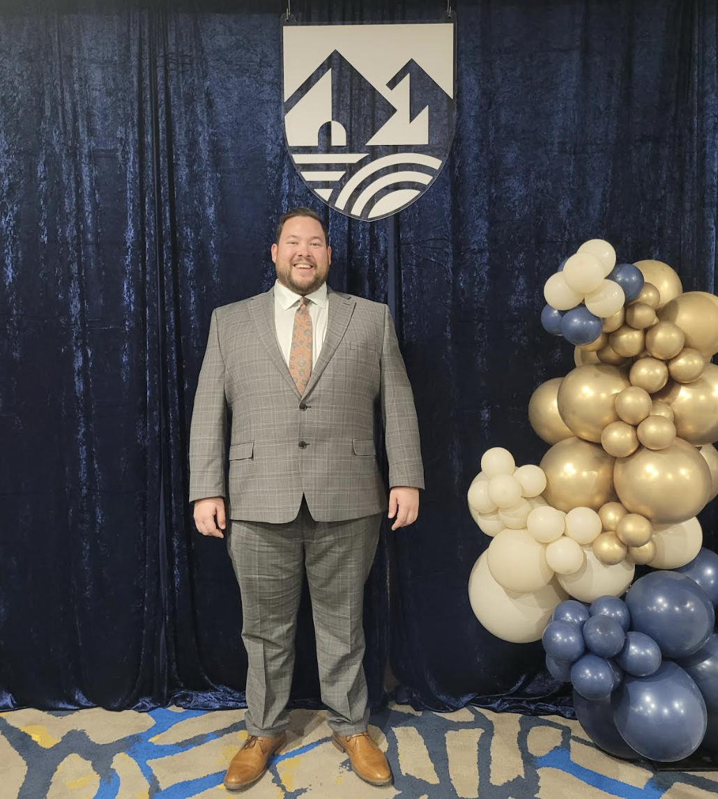 A formal portrait of Christopher Clouthier standing and smiling in front of a dark blue velvet curtain at an event. He is wearing a grey plaid suit, a white dress shirt, a patterned tie, and brown dress shoes. To his left stands a large, decorative vertical cluster of white, gold, and dark blue balloons. 