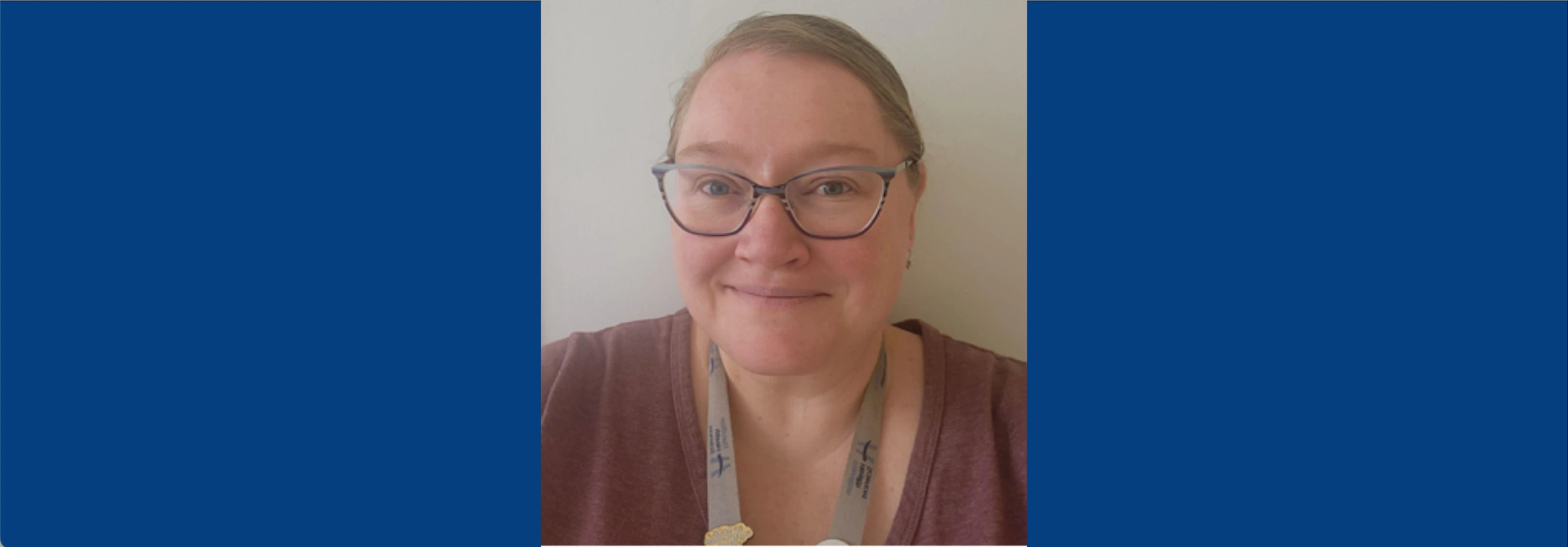 A close-up portrait of a smiling Sarah Ferguson. She has light brown hair pulled back, wears striped glasses, and is dressed in a mauve t-shirt with a grey Hamilton Health Sciences lanyard.