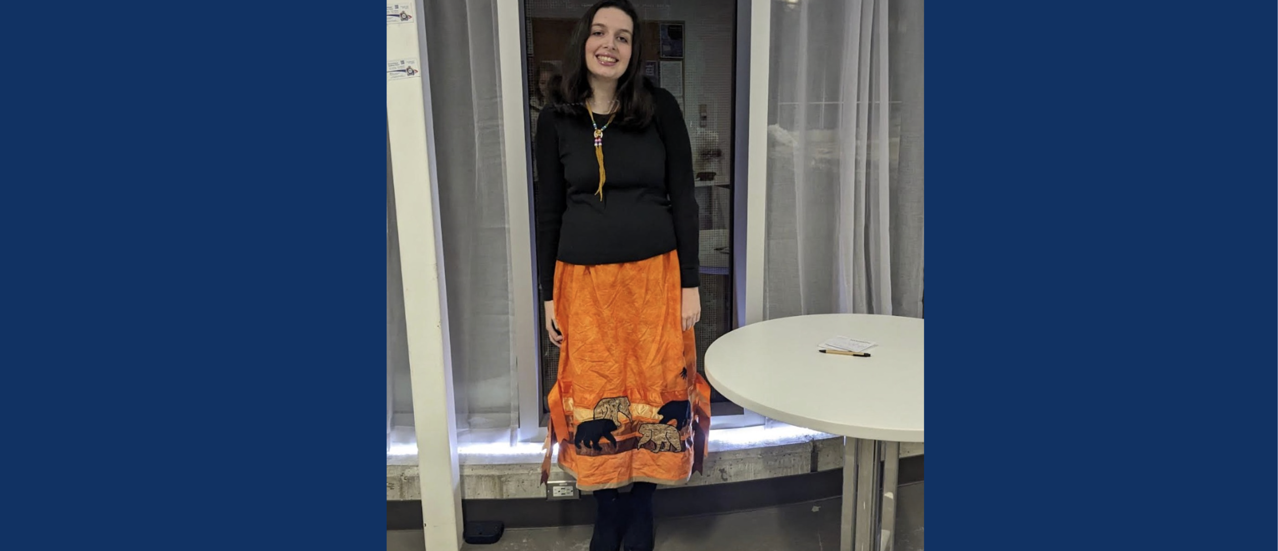 A portrait of Kailey O'Bumsawin smiling while standing indoors next to a round white table. She is wearing a long-sleeved black shirt, a traditional beaded necklace, and a bright orange ribbon skirt adorned with black and tan bear silhouettes along the bottom hem.