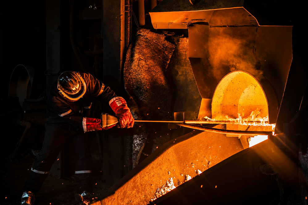 Worker in protective gear operating a high-temperature industrial furnace, illuminated by the glow of molten material.