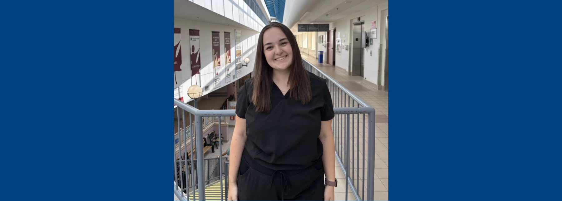 Emma Bélanger smiling in black scrubs on an indoor hospital balcony under a bright glass skylight.