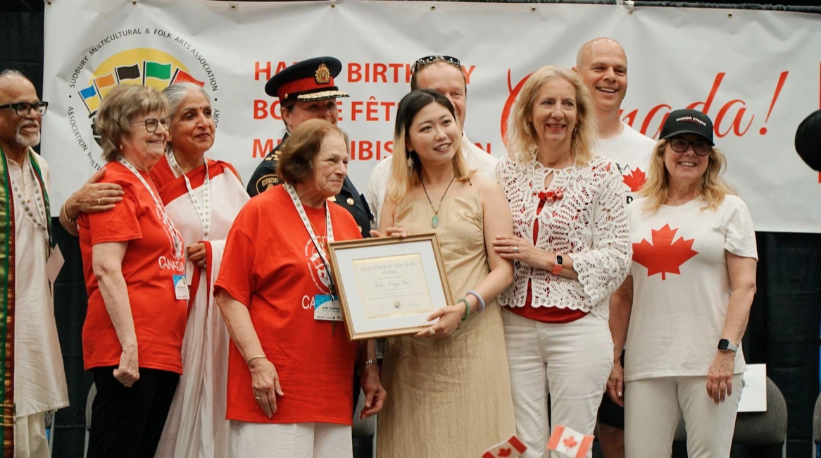 Felice Guo standing proudly on stage at the Greater Sudbury Canada Day celebrations, smiling and holding her framed "Newcomer of the Year" award certificate. She is surrounded by diverse community members, event organizers, and local officials, some wearing red and white in front of a celebratory banner.