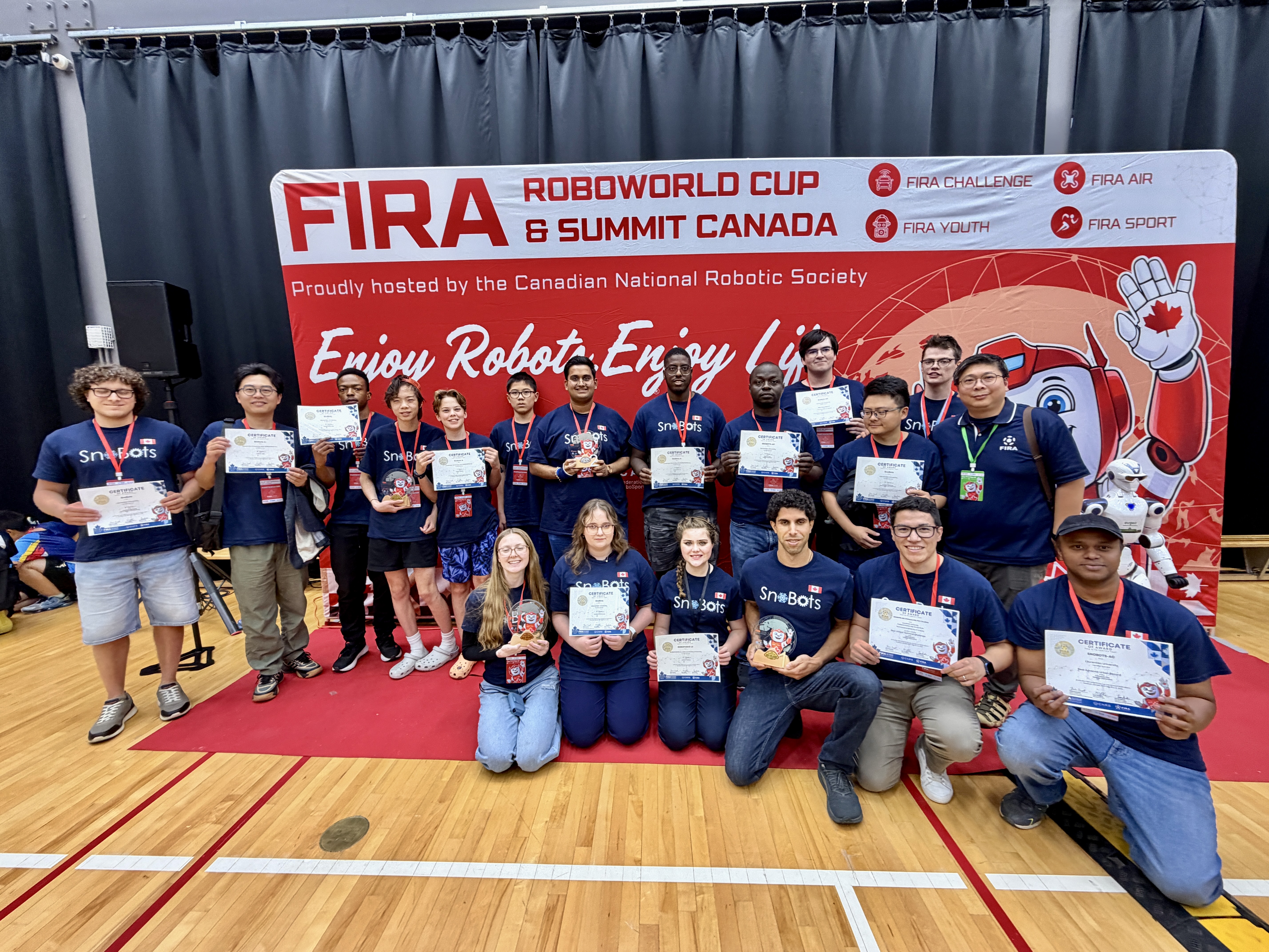Members of the Laurentian University SnoBots robotics team and youth competitors pose on a red carpet with their certificates and trophies in front of a FIRA RoboWorld Cup Canada banner.