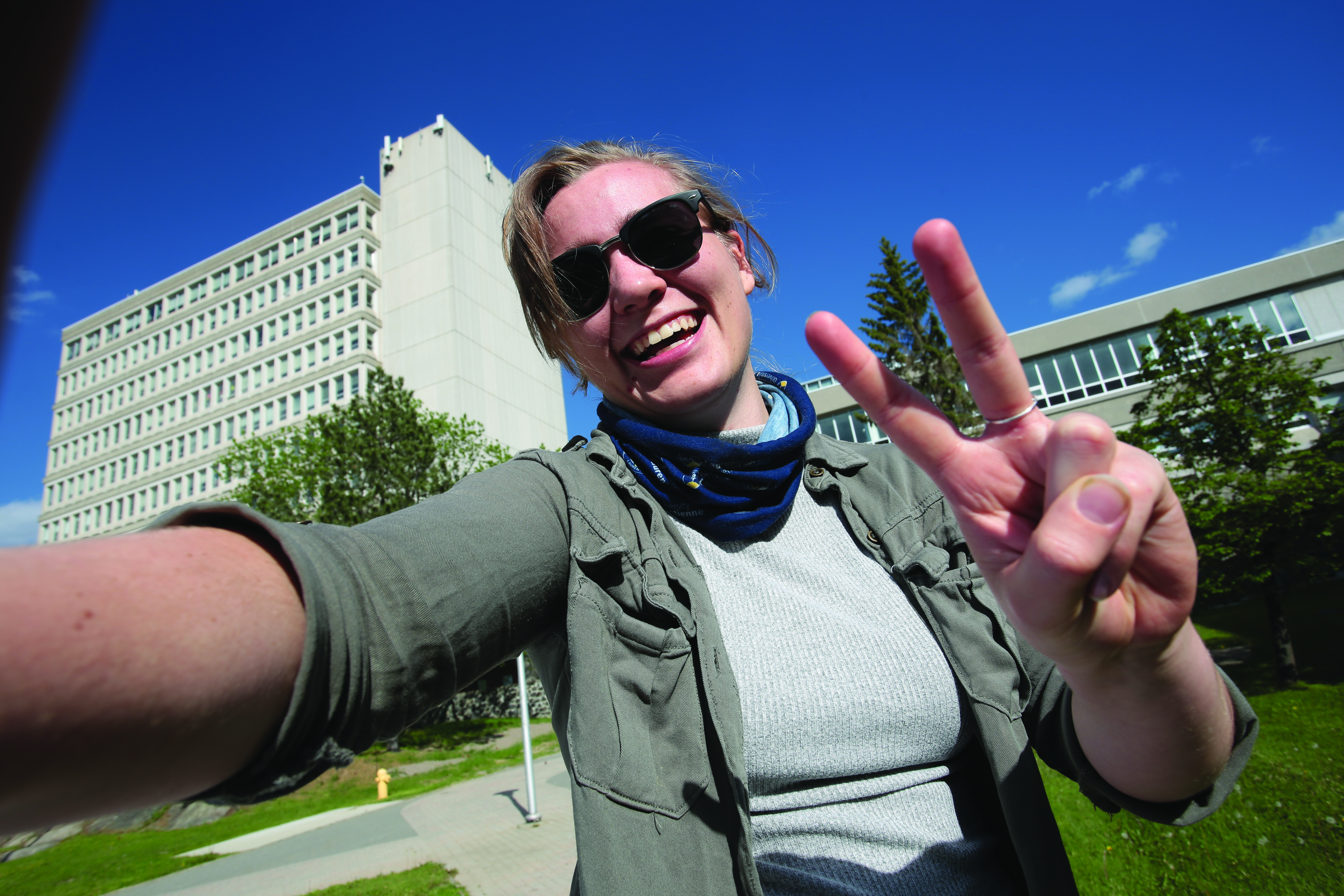 A smiling student outdoors on campus taking a selfie and making a peace sign, with campus buildings in the background.