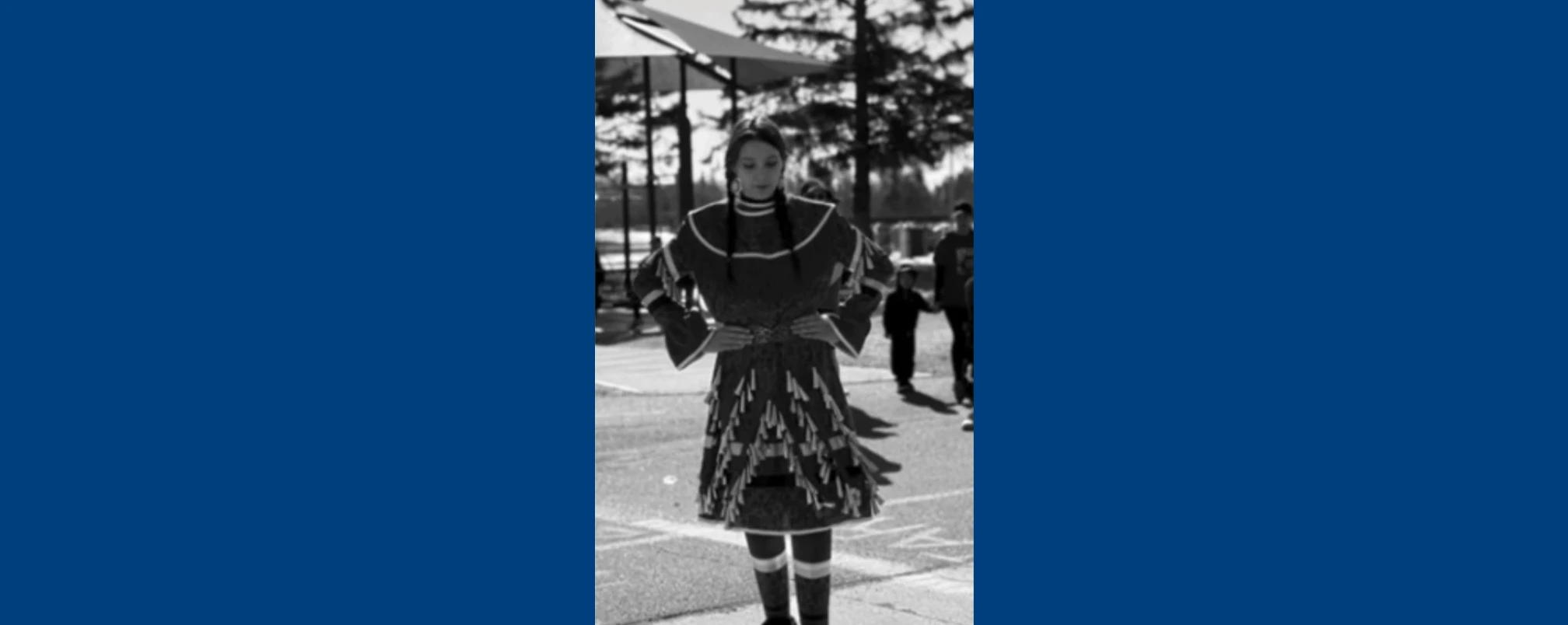 Black and white photograph of Kimberly Kennedy standing outdoors in a traditional jingle dress with her hands on her hips, looking down. In the background, trees and community members are visible.
