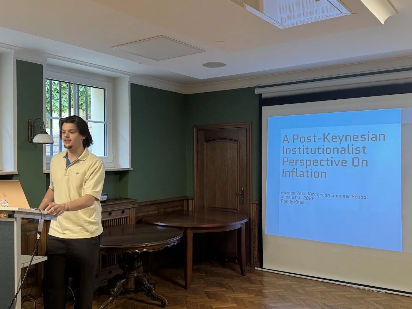 Randy Kneer standing at a podium delivering an economic lecture in a classroom in Poland. Next to him, a large projector screen displays his presentation title slide, reading "A Post-Keynesian Institutionalist Perspective On Inflation" by Randy Kneer, dated June 21st, 2026.