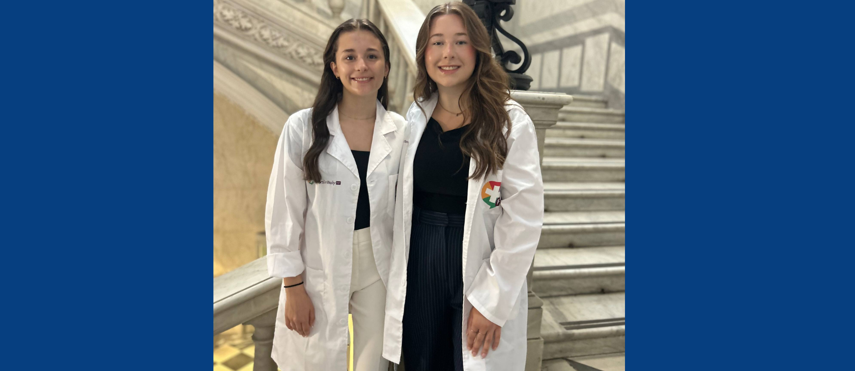 Medical fellowship students Karina Schryer and Kaleigh Hamilton smiling side-by-side in white lab coats on a grand marble staircase inside the Azienda Ospedaliero-Universitaria in Alessandria, Italy.