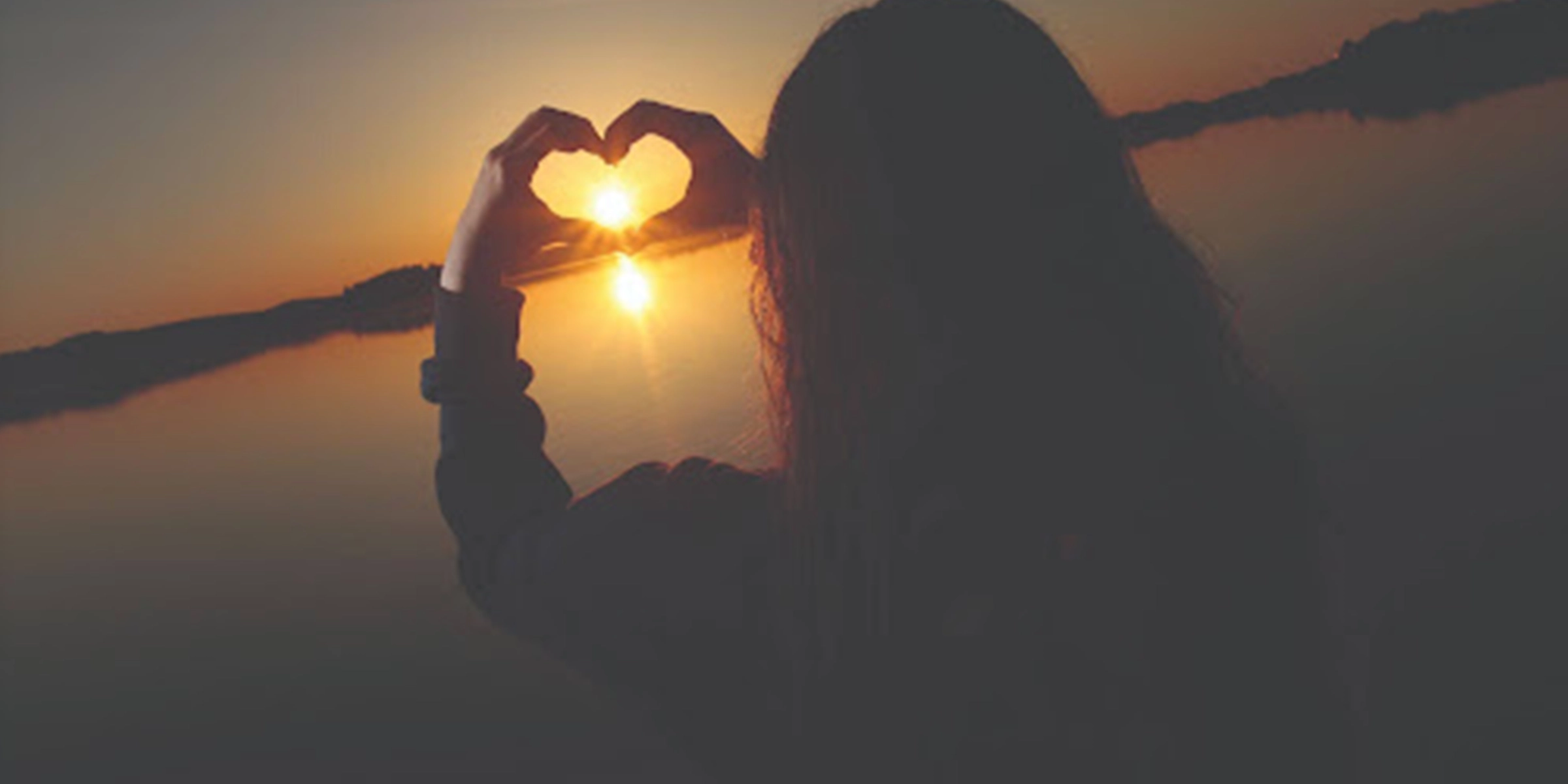 Person making a heart with her hands towards a sunset