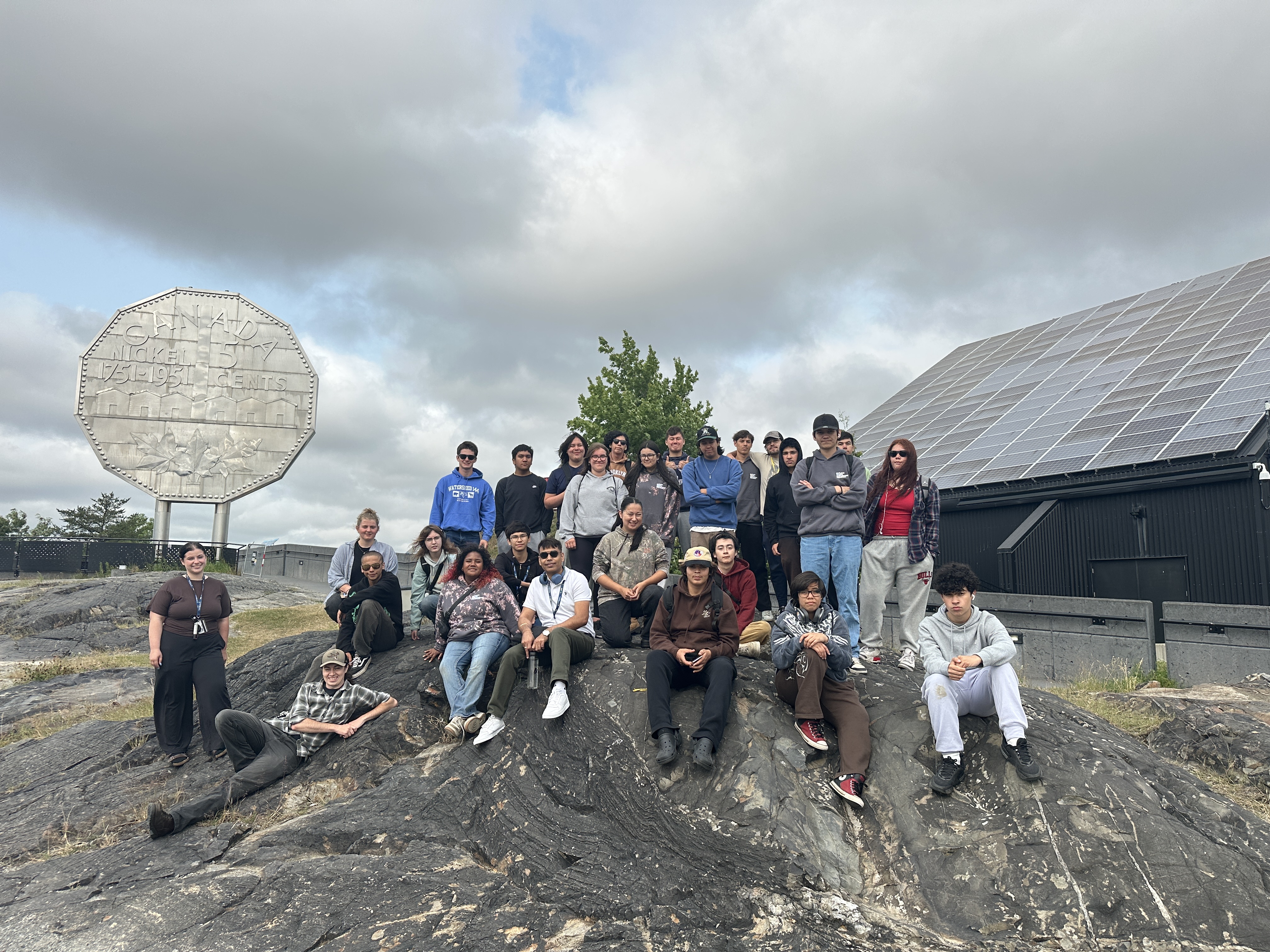 OYEP participants and staff posing outdoors on a rocky hill in front of the Big Nickel monument and solar panels at Dynamic Earth. 