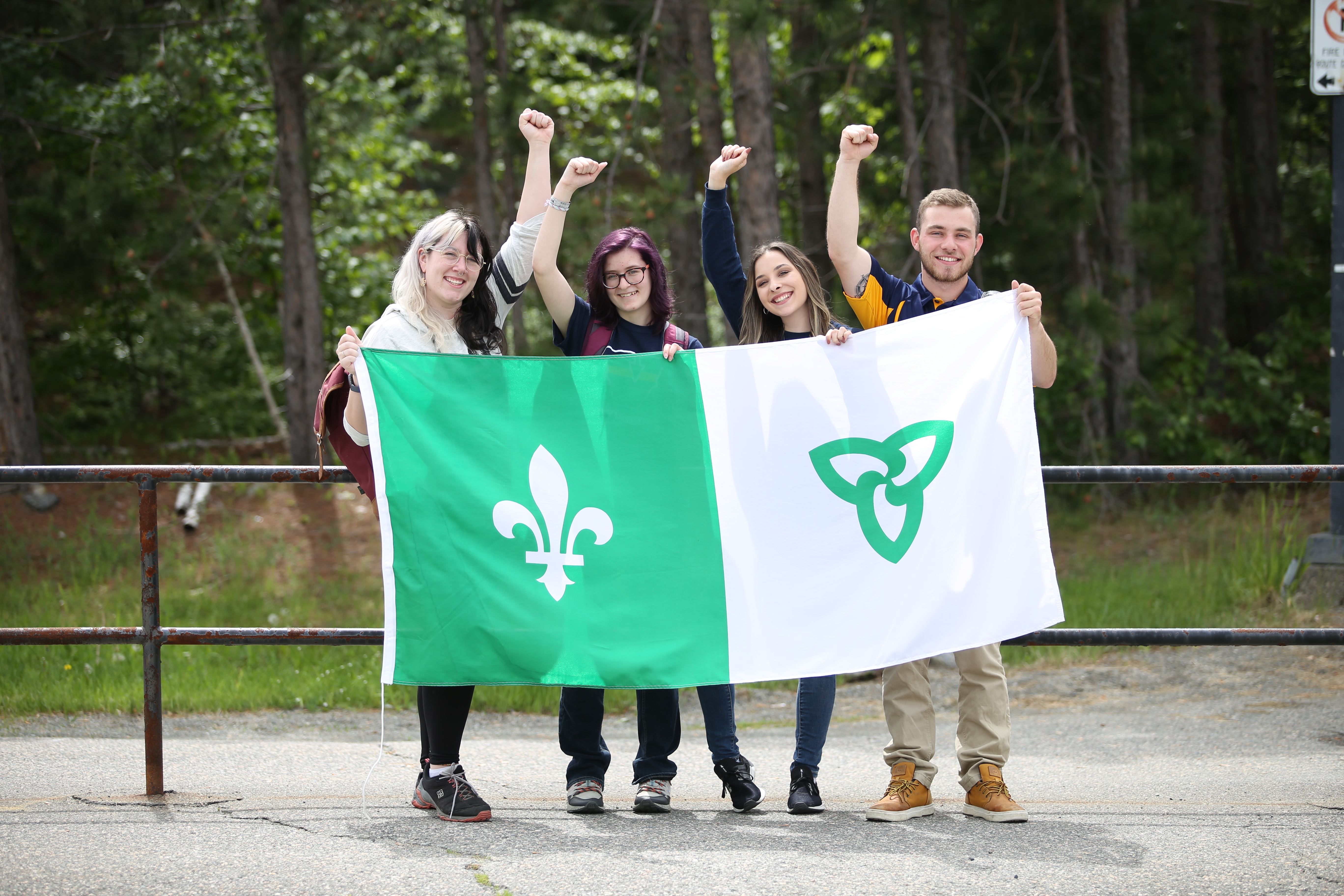 Four students standing outdoors holding a large Franco-Ontarian flag with raised fists, smiling in front of a wooded area.