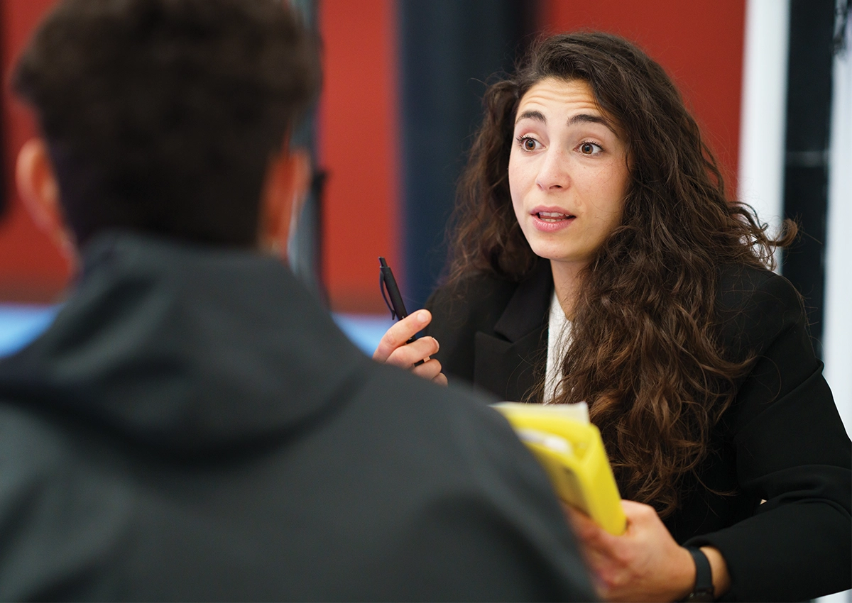 A lady with a pen and clipboard speaking to someone out of focus in the foreground
