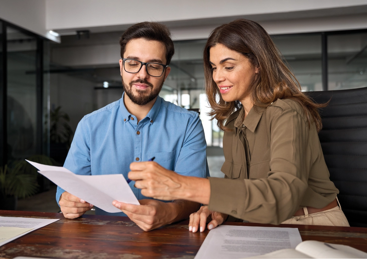 Two people sitting at a desk with a woman showing the man something on a piece of paper.