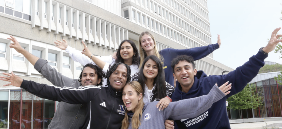 A diverse group of university students smiling and posing together outdoors in front of the Parker Building at Laurentian University, with several extending their arms outward enthusiastically.