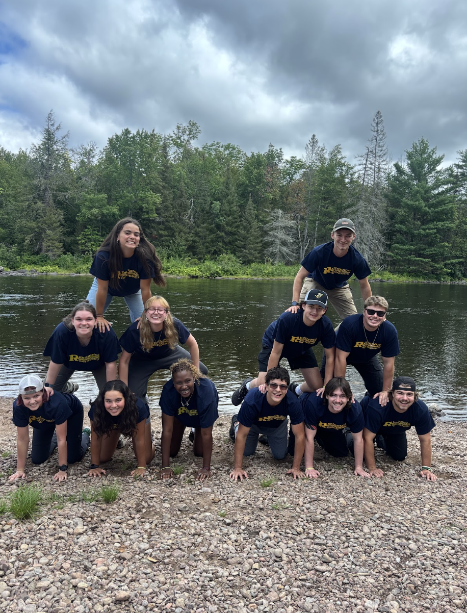 A group of eleven Residence Assistants wearing matching dark navy "Residence Staff" t-shirts pose in two human pyramids on a rocky shoreline in front of a river and forest.