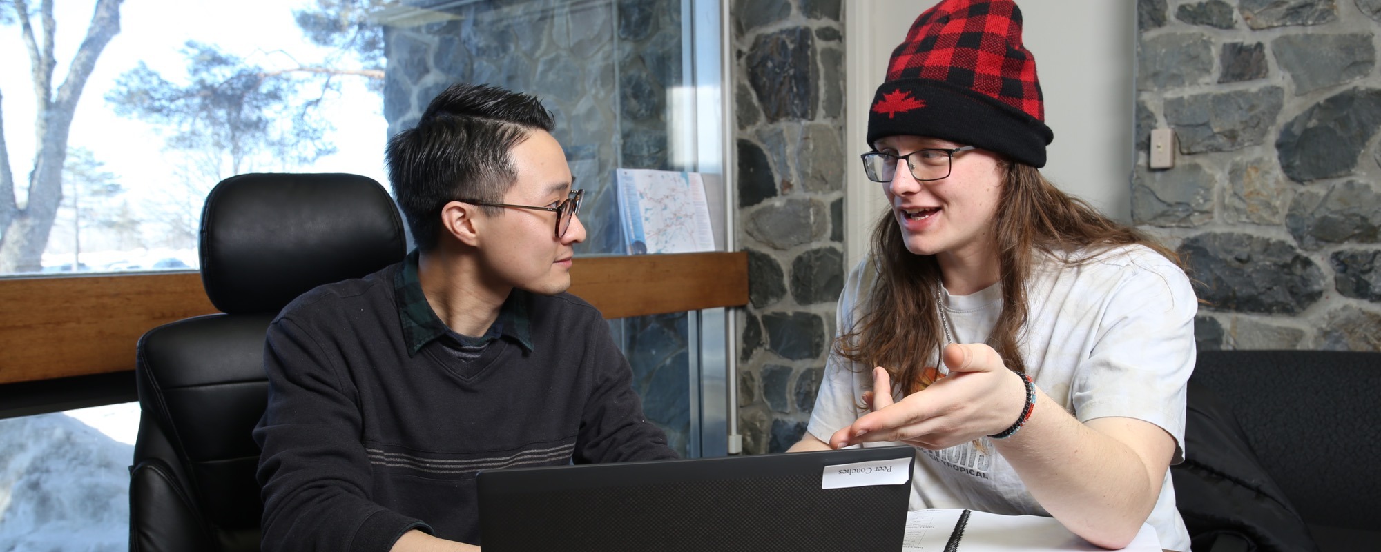Deux étudiants assis à un bureau devant un ordinateur qui se parlent.