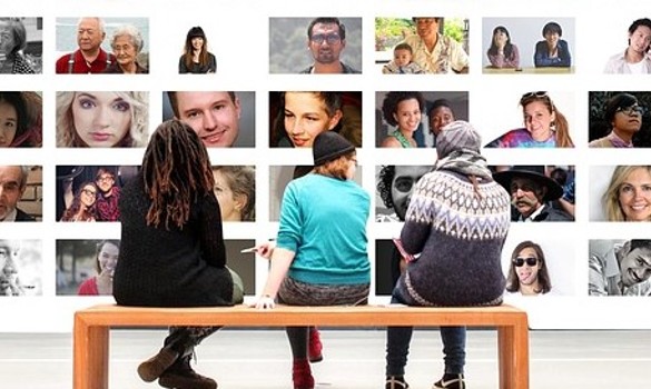 Three people sitting at a bench looking at a series of images on a wall of different people
