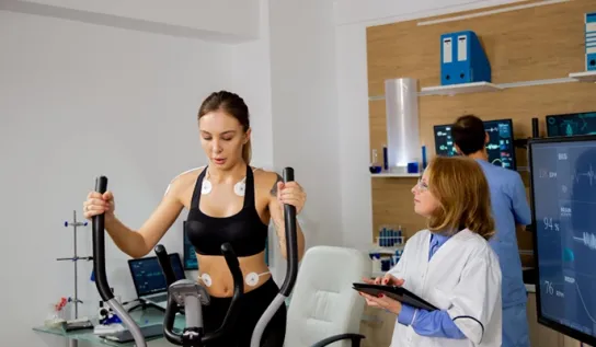 A woman on an elliptical with vital monitoring equipment on her and a researcher observing her.