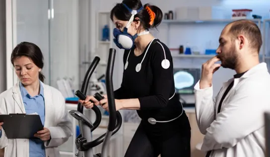 A woman on an elliptical with electrodes and a respiration monitoring mask on her, and with two researchers standing on each side of her monitoring her.