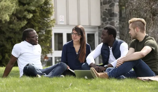 Four students sit on a field of grass, talking to each other.