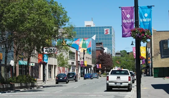 A downtown Sudbury street, with cars parked on the side and a building with a "you are beautiful" mural.