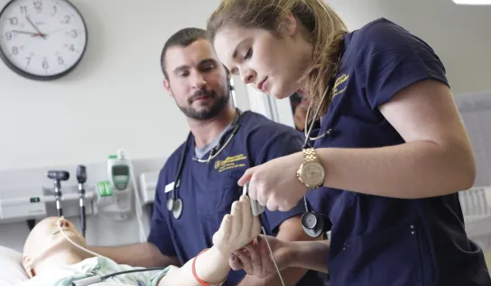 Nursing students applying a pulse monitor on the hand of a medical manikin.