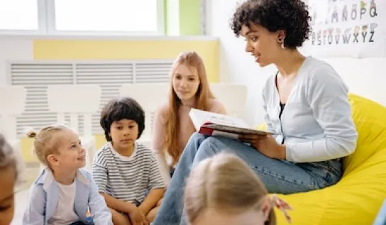 A teacher sitting and reading a book to her students who are sitting around her.