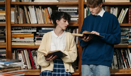 Two students standing next to each other and reading books in a library.