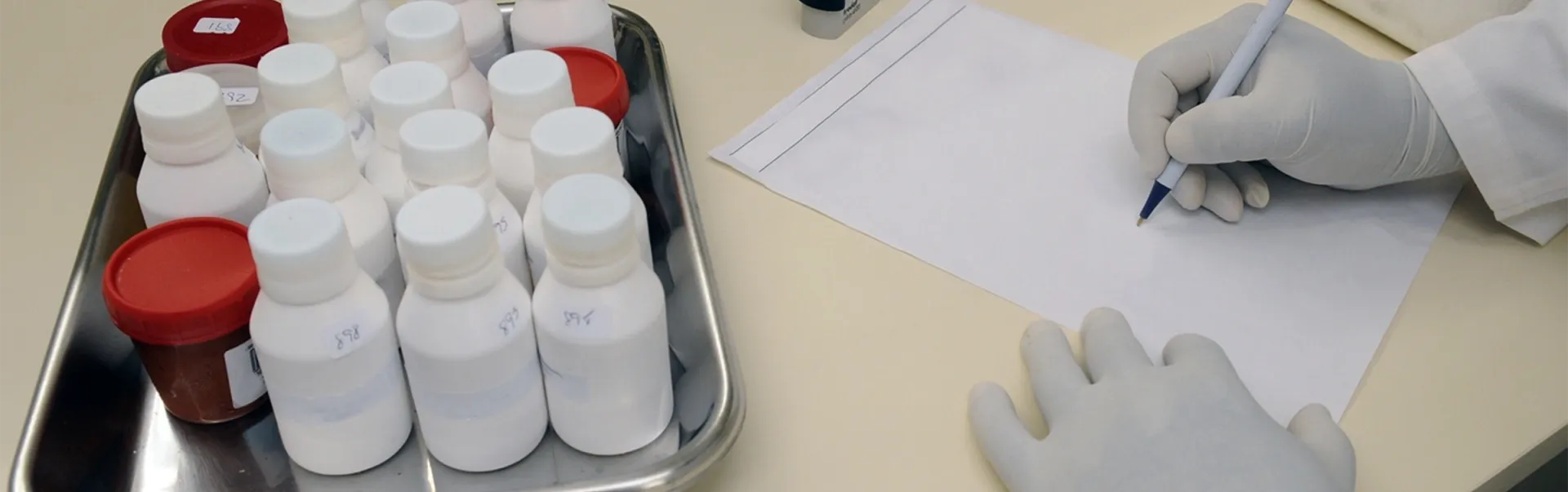 A laboratory worker writing on a sheet of paper, with a tray of bottles next to them.