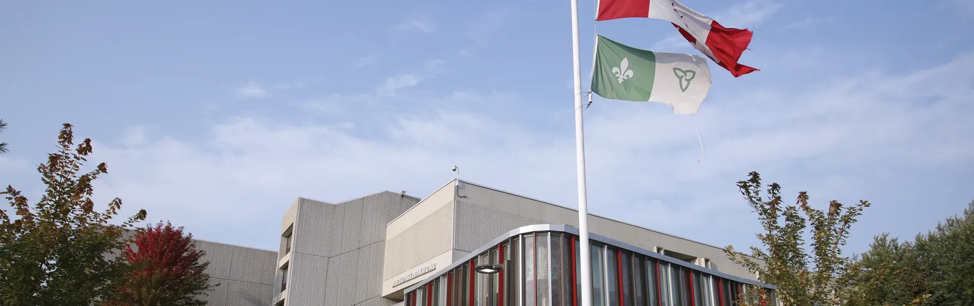 The Alphonse Raymond building with the Canadian and Franco-Ontarian flags flying on the flagpole outside.